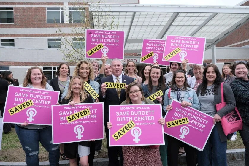 A group of women and one man stand holding "Save Burdett Birth Center" signs.